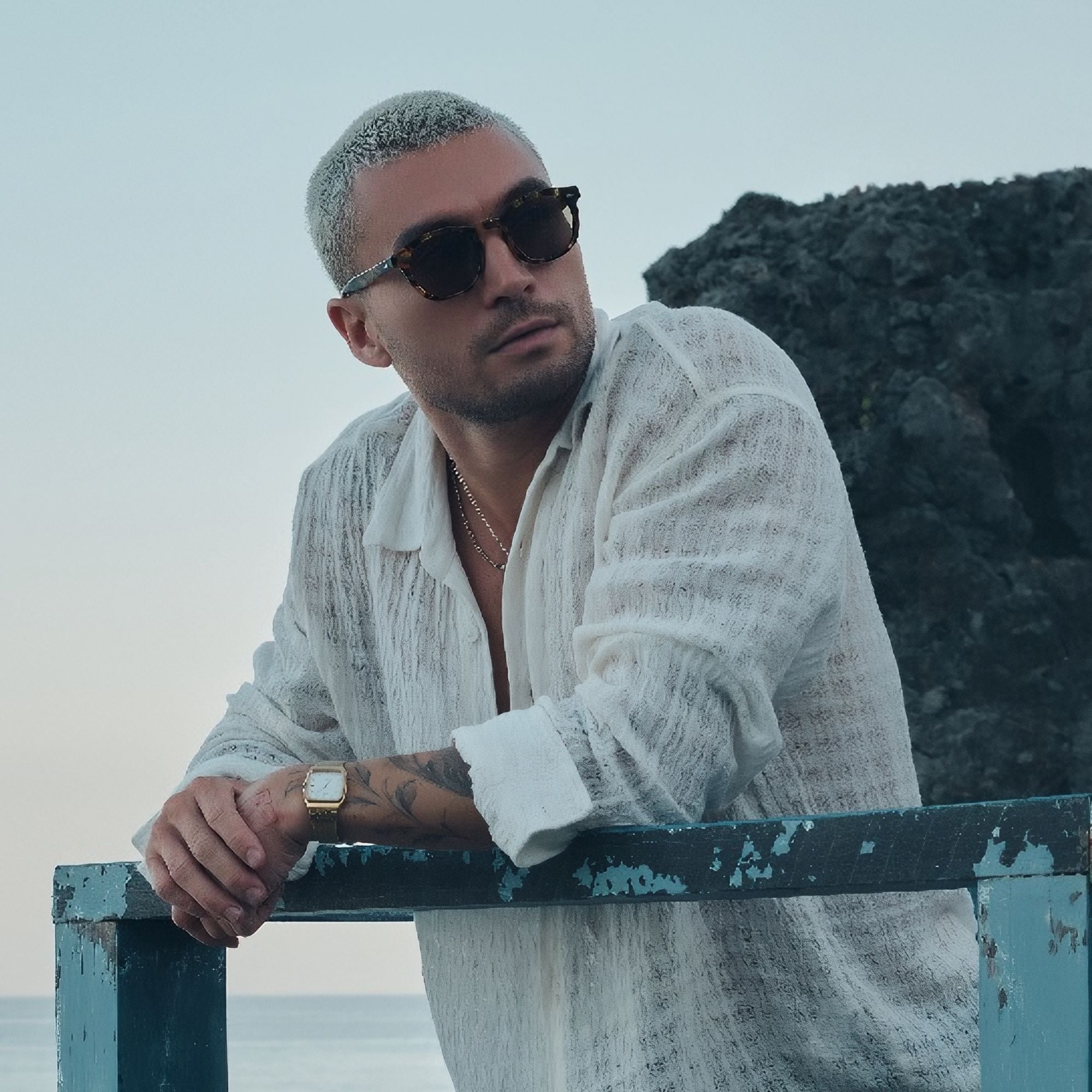 Man wearing sunglasses and a white shirt leaning on a railing with a scenic background